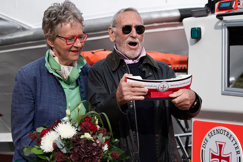 Ältere Dame hält vor einem neuen Seenotrettungsboot einen Blumenstrauß in der Hand, und ein älterer Herr hält ein Seenotretter-Sammelschiffchen in der Hand