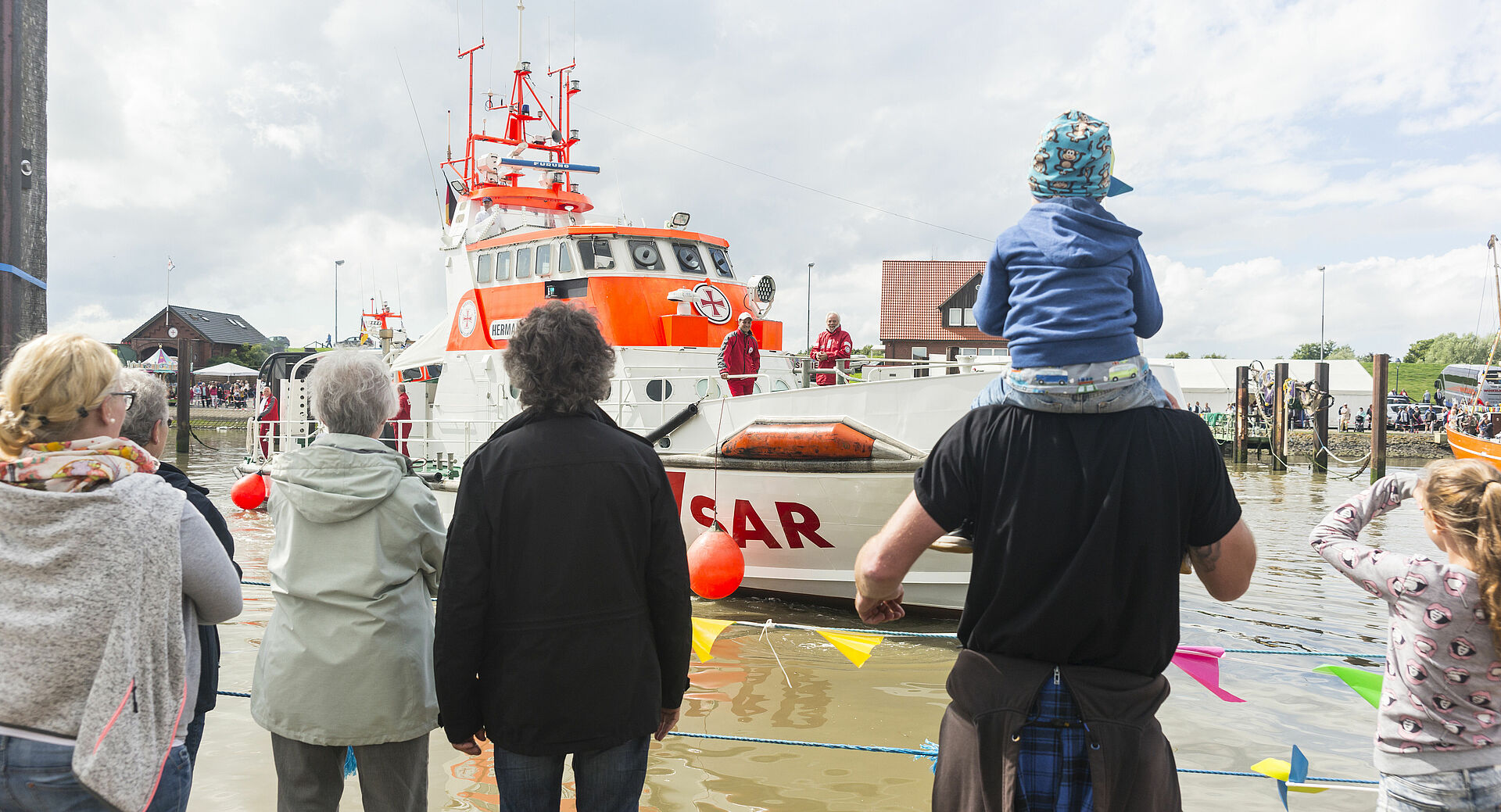 Blick von hinten auf eine Gruppe von Touristinnen und Touristen, die auf einen Seenotrettungskreuzer schauen, der im Hafen von Fedderwardersiel anlegt.