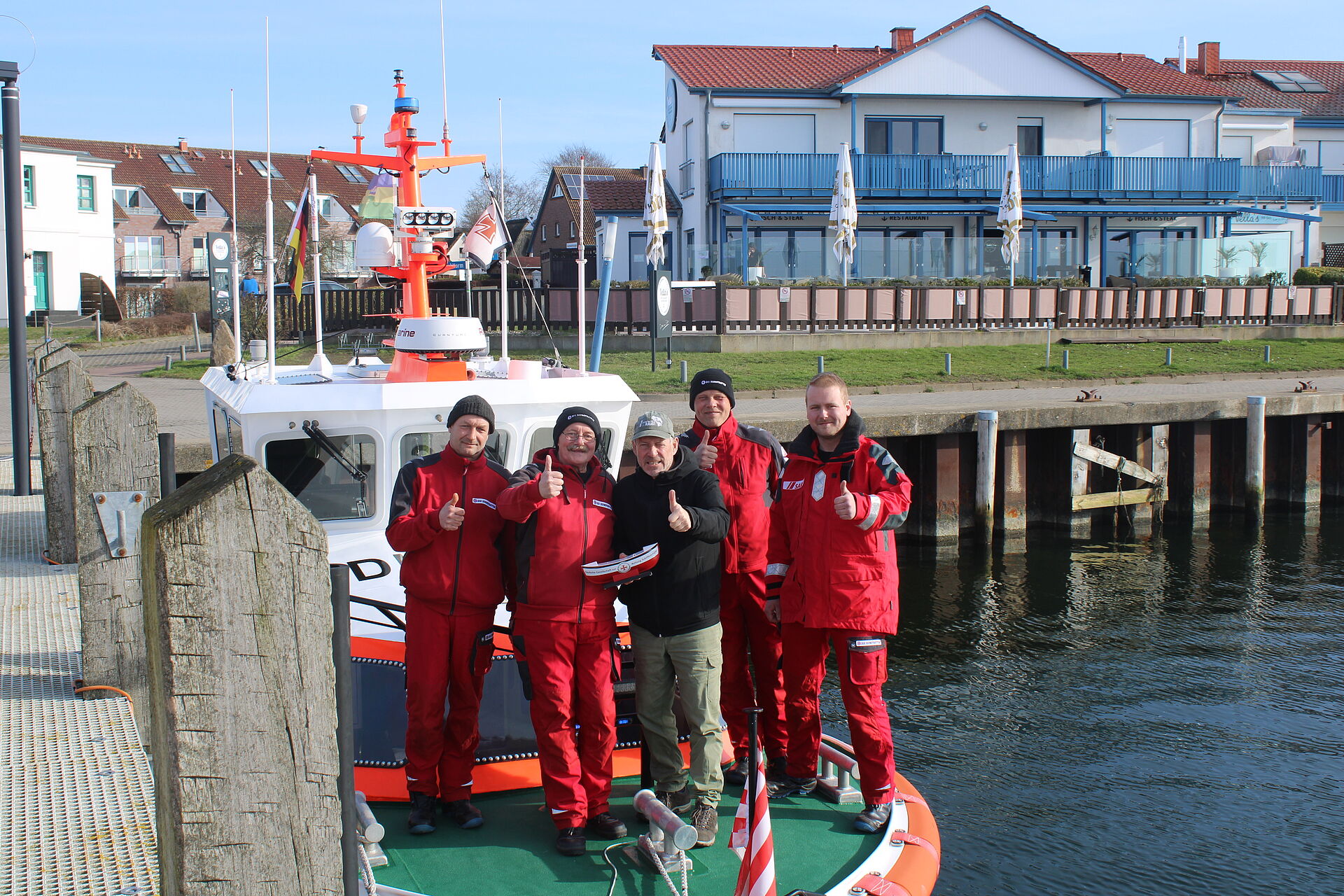 Spendenübergabe auf einem Seenotrettungsboot: Auf diesem stehen vier freiwillige Seenotretter in roter Dienstkleidung. Sie rahmen den NDR-Moderator ein, der ein DGzRS-Sammelschiffchen in den Händen hält. Alle halten einen Daumen hoch.