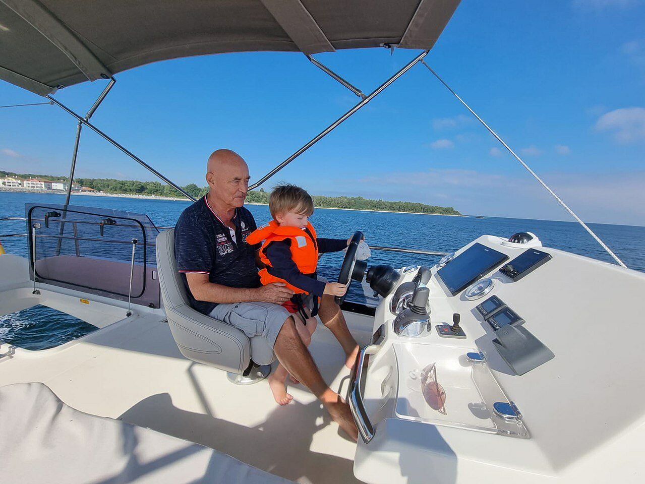 Der Verstorbene mit einem seiner Enkel am Ruder seines Motorbootes auf dem Wasser, bei blauem Himmel, im Hintergrund ist noch das Festland zu sehen.