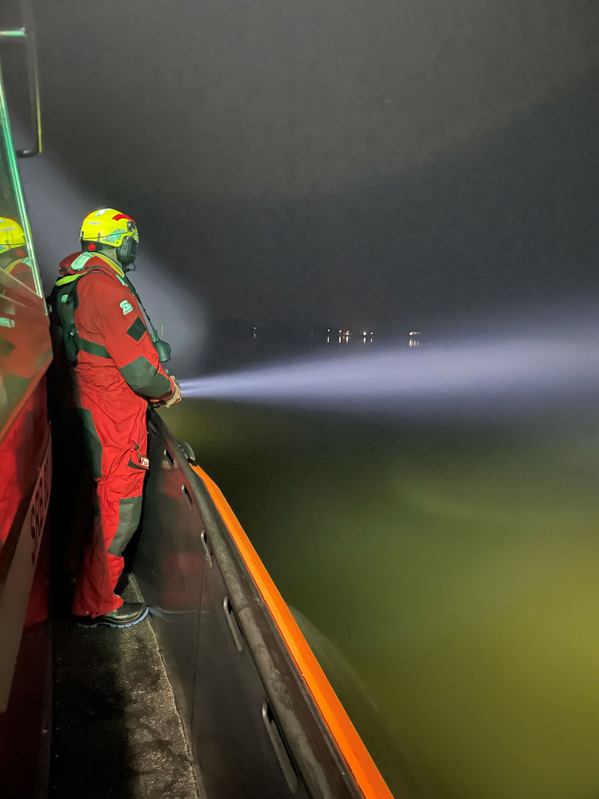 Seenotretter steht bei nächtlichem Einsatz an der Reling und leuchtet mit Taschenlampe auf das Wasser
