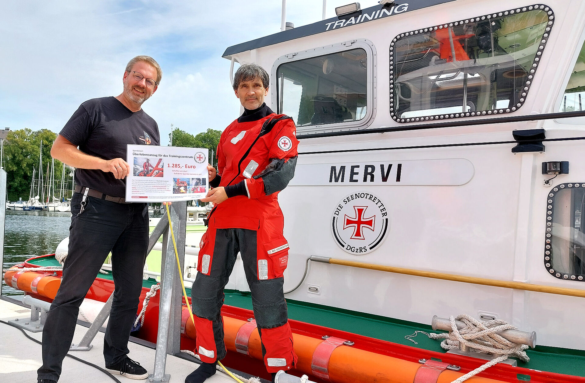 Vor dem Trainingsboot MERVI stehen der Liedermacher (l.) aus Kehl am Rhein und der Leiter des DGzRS-Trainingszentrums, im roten DGzRS-Kälteschutzanzug, mit einem symbolischen Spendenscheck in den Händen.