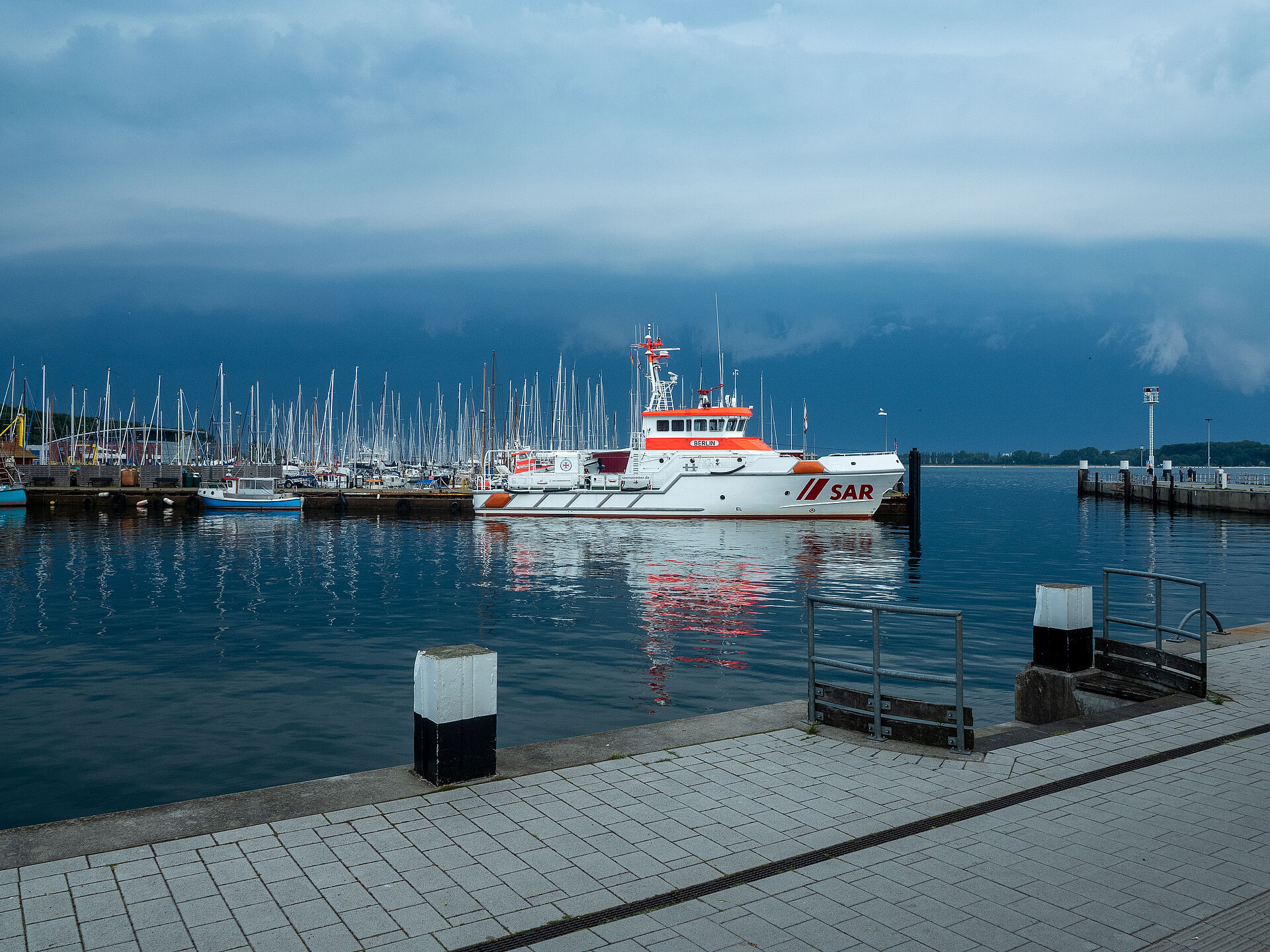 Der Seenotrettungskreuzer BERLIN liegt an seinem Liegeplatz im Hafen von Laboe und spiegelt sich im Hafenbecken. Dunkelblaue Regenwolken schweben über der Szenerie.