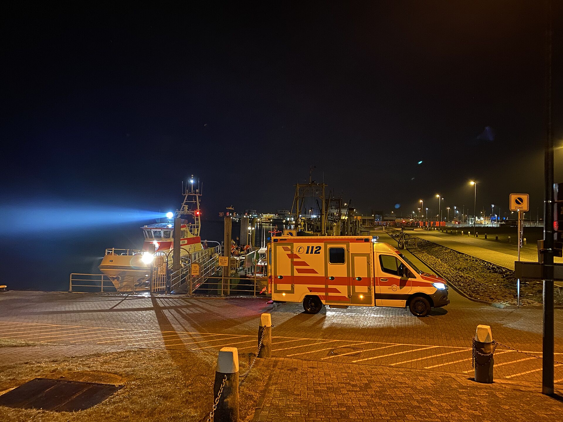 Nachtszene im Hafen: Im Vordergrund am Anleger steht ein Rettungswagen, dahinter hat ein Seenotreettungskreuzer festgemacht. Seine Scheinwerfer leuchten in den dunklen Himmel über dem Hafengelände.
