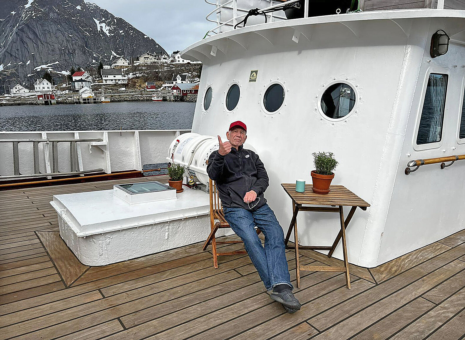 Ein Mann sitzt an Bord eines Schiffes auf einem Klappstuhl; im Hintergrund ist ein Hafen und schneebeckte Berge zu sehen.