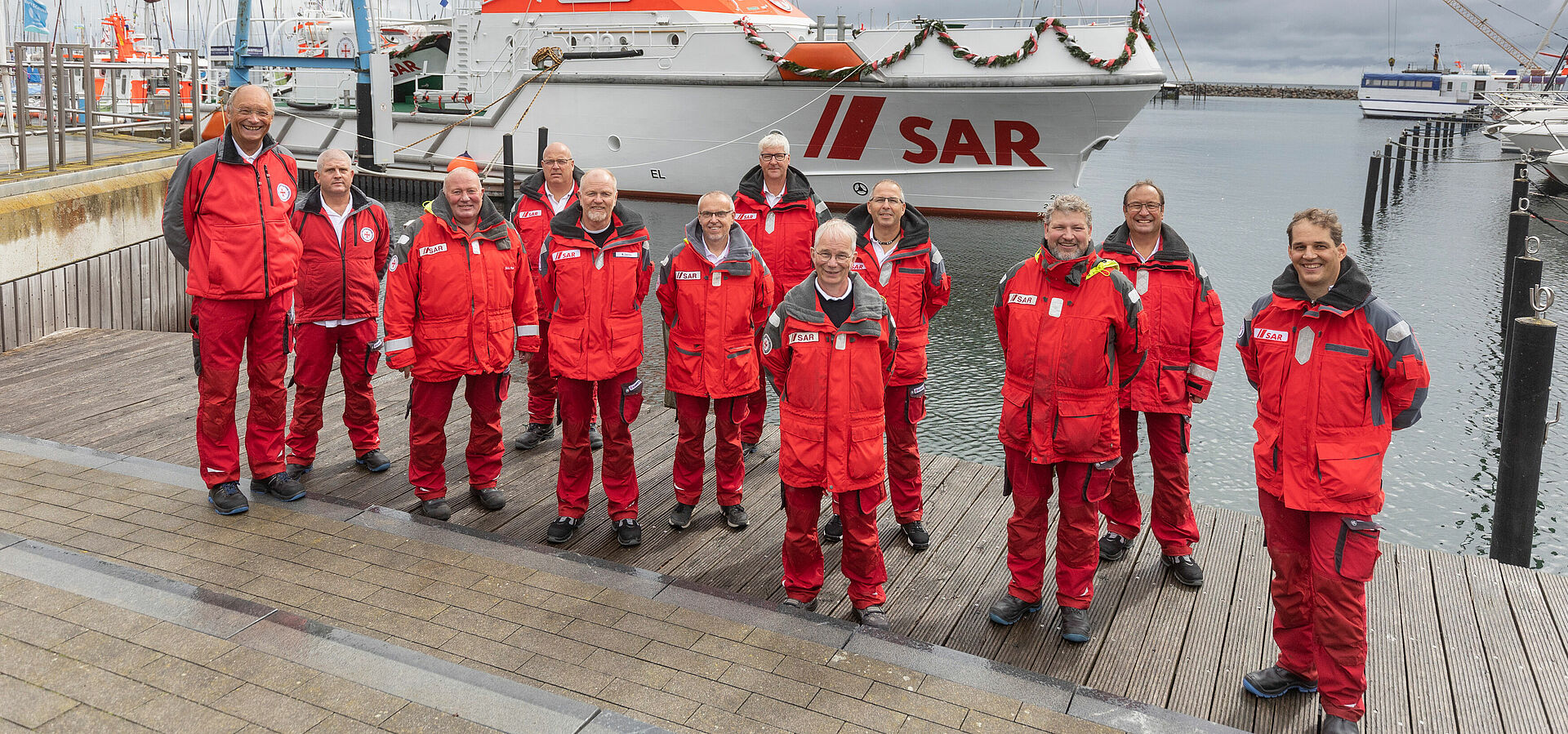 Gruppenbild der Crew von Grömitz. Sie stehen in roter Einsatzkleidung, jeweils mit etwas Abstand, an der Hafenkante im Grömitzer Hafen vor dem Seenotrettungskreuzer FELIX SAND.