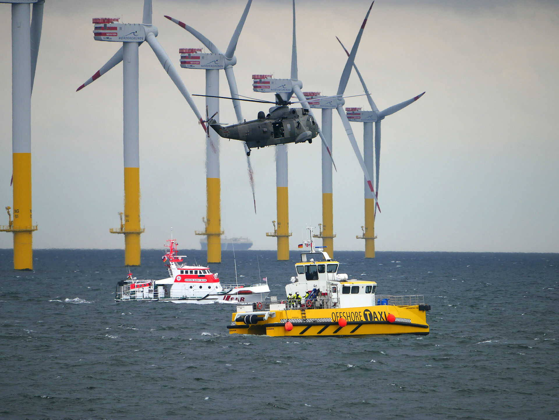 Blick auf einen Offshore-Windpark mit DGzRS-Seenotrettungskreuzer, SAR-Hubschrauber der Marine und einem Offshore-Taxi im Vordergrund vor sechs Windrädern.