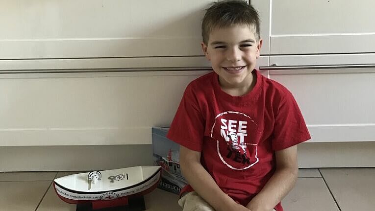 Junge im roten Seenotretter-T-Shirt sitzt auf dem Küchenfußboden, neben ihm steht ein Sammelschiffchen. Hinter ihm auf dem Schrank stehen Lego-Fahrzeuge und hängen selbstgemalte Bilder von Seenotrettungskreuzern.