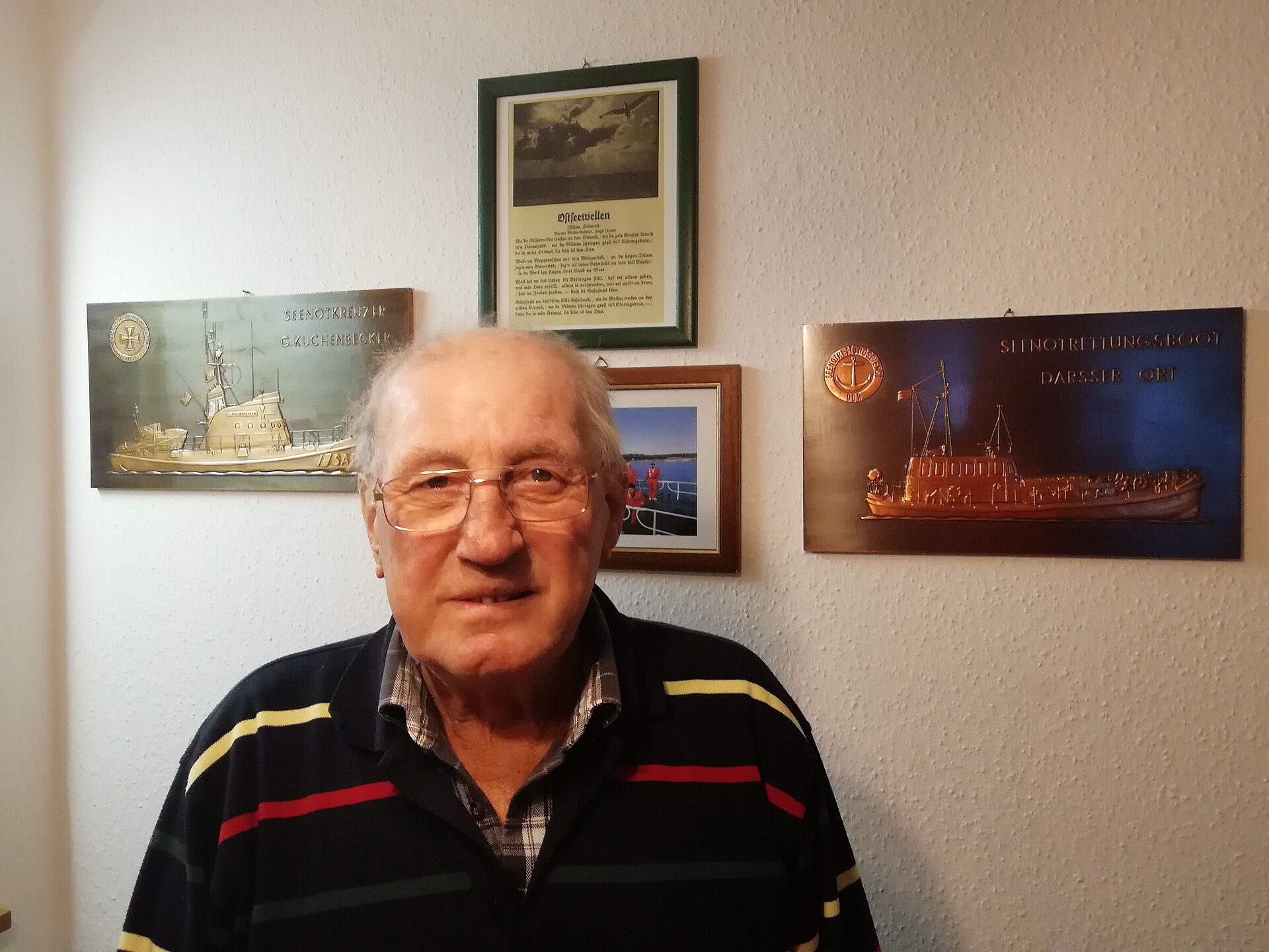 Foto von einem alten Mann vor einer Wand an der Bilder von Schiffen hängen
