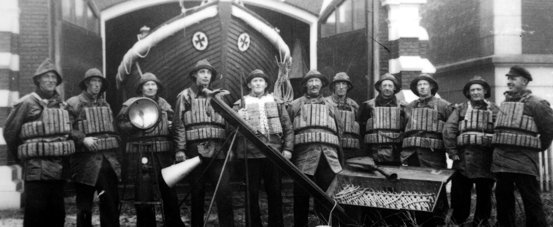 Seenotretter stehen mit historischem Ölzeug und Kork-Schwimmwesten vor einem Ruderrettungsboot