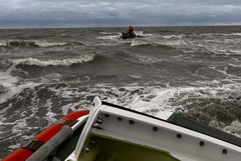 Das Heck eines Seenotrettungsbootes, dahinter in der Brandung ein Wrack mit zwei Menschen darauf. Zur Seite: Marineflieger retten Schiffbrüchige aus Seenot