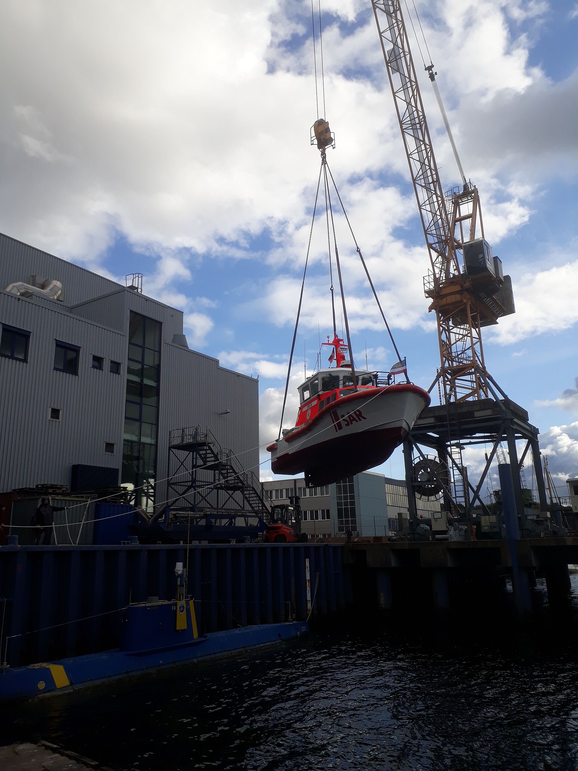 Ein großer Hafenkran hebt das Seenotrettungsboot vom Hof in das Hafenbecken der Werft.