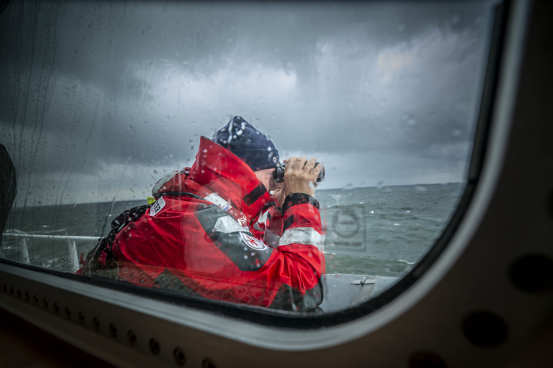 Bei regnereischem, schlechten Wetter steht ein Seenotretter mit Mütze auf einem Ausguck und blickt mit einem Fernglas über die aufgewühlte See.