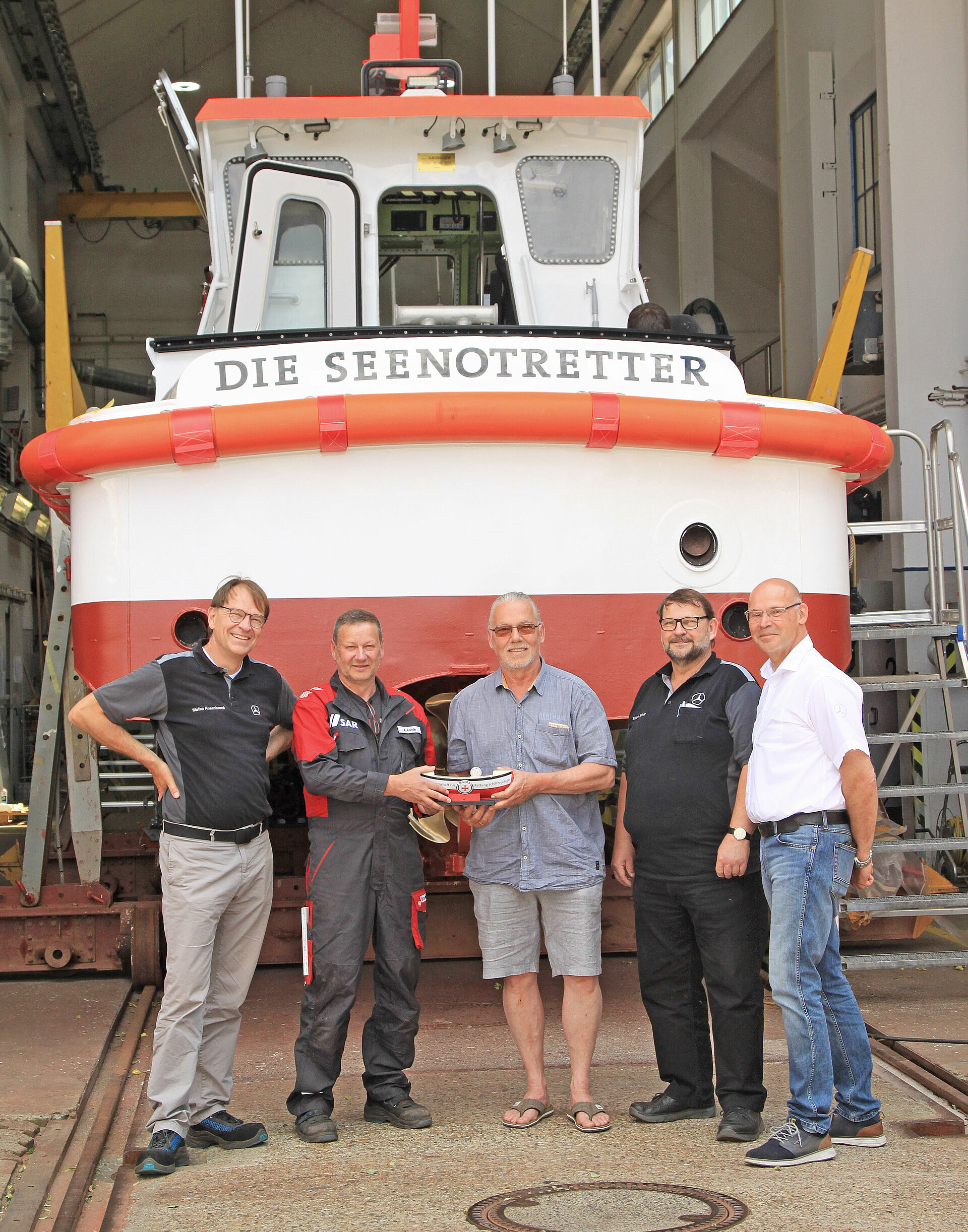 Fünf Männer stehen vor einer Werfthalle, in der ein Seenotrettungsboot zu sehen ist.