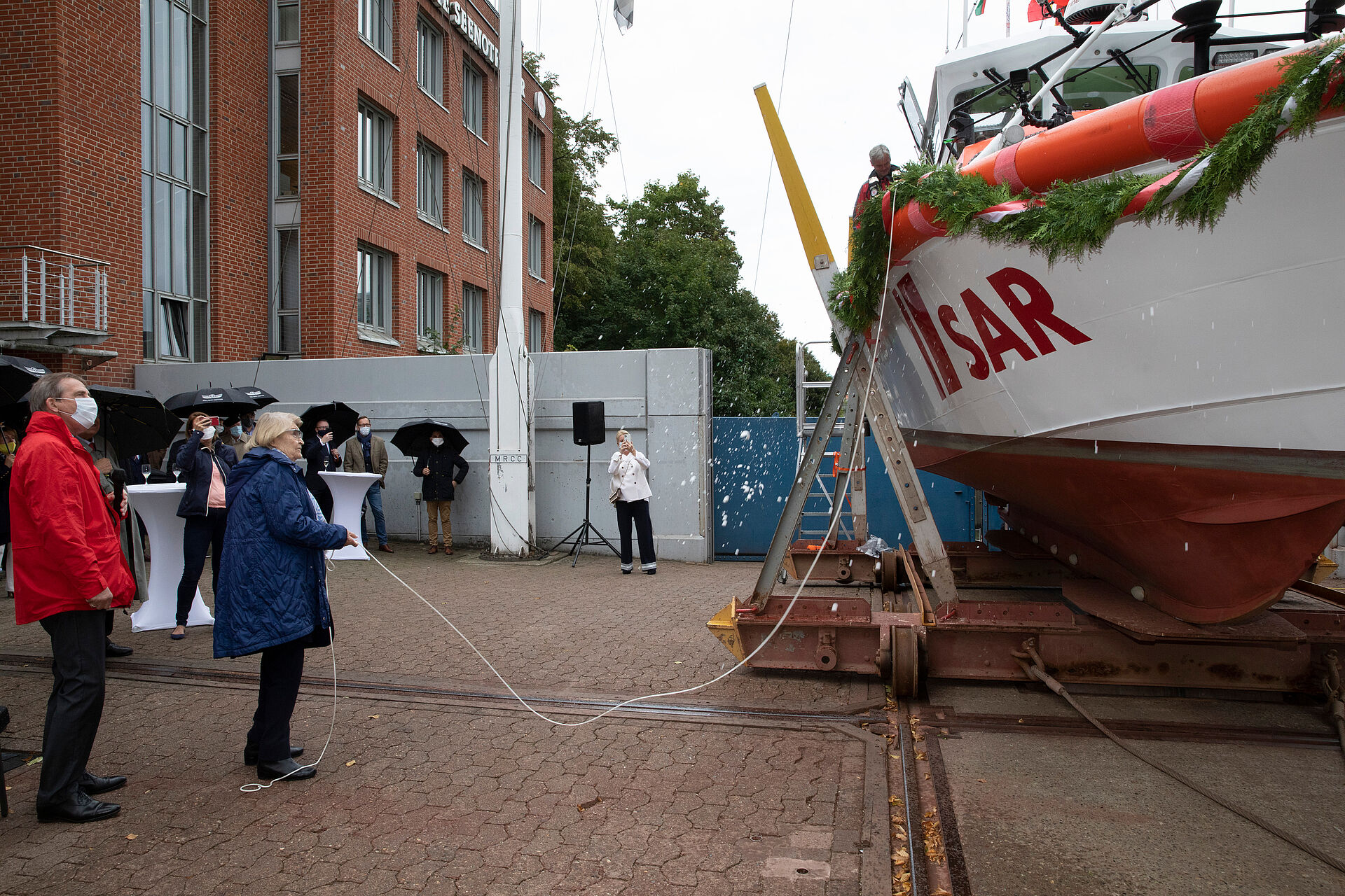 Vor dem geschmückten Seenotrettungsboot auf dem Hof der DGzRS-Zentrale steht die Taufpatin in blauer Jacke und hält das Seil in der Hand, das mit der Sektflasche an einer Stange, die am Boot befestigt ist, verbunden ist.