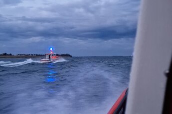 Von einem fahrenden Seenotrettungsboot ist ein anderes rot-weißes Seenotrettungsboot auf See aufgenommen worden. Es hat Blaulicht an. Dunkle Wolken sind am Himmel, es wird langsam dunkel.. Zur Seite: Seenotretter befreien zwei Kiter aus Notlage