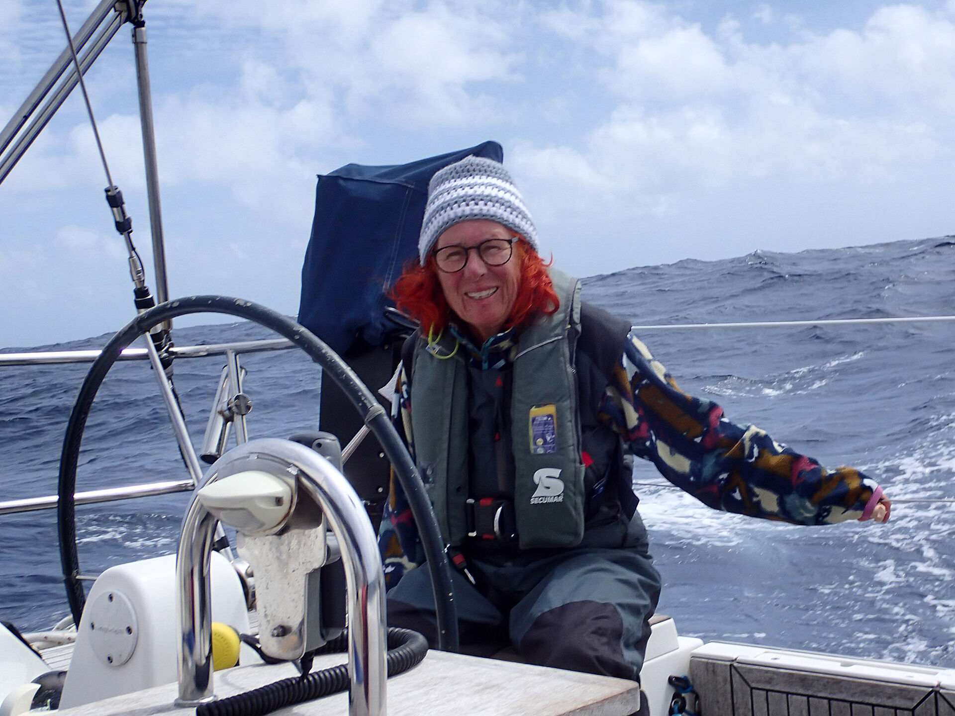 Seglerin und Spenderin Brigitte Honnens sitzt an Bord Ihrer Segelyacht, bekleidet mit Rettungsweste und Pudelmütze. Im Hintergrund Wasser und blauer Himmel, vor ihr das Ruder.