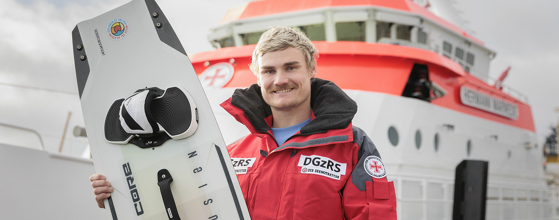 Linus Erdmann steht mit roter Wetterjacke vor einem Seenotrettungskreuzer und hält sein Kiteboard.