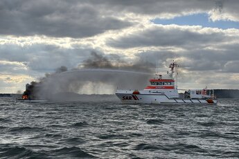 Qual steigt von einem brennendem Boot auf. Ein rot-weißer Seenotrettungskreuzer löscht es.. Zur Seite: Segelyacht vor Travemünde in Flammen – Motorboot-Crew rettet Skipper