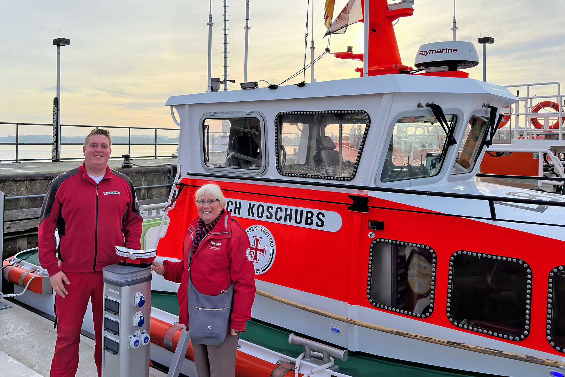 Vormann Patrick Morgenroth und die ehrenamtliche Mitarbeiterin Petra Petersen vor dem Seenotrettungsboot ERICH KOSCHUBS mit einem DGzRS-Sammelschiffchen vor sich