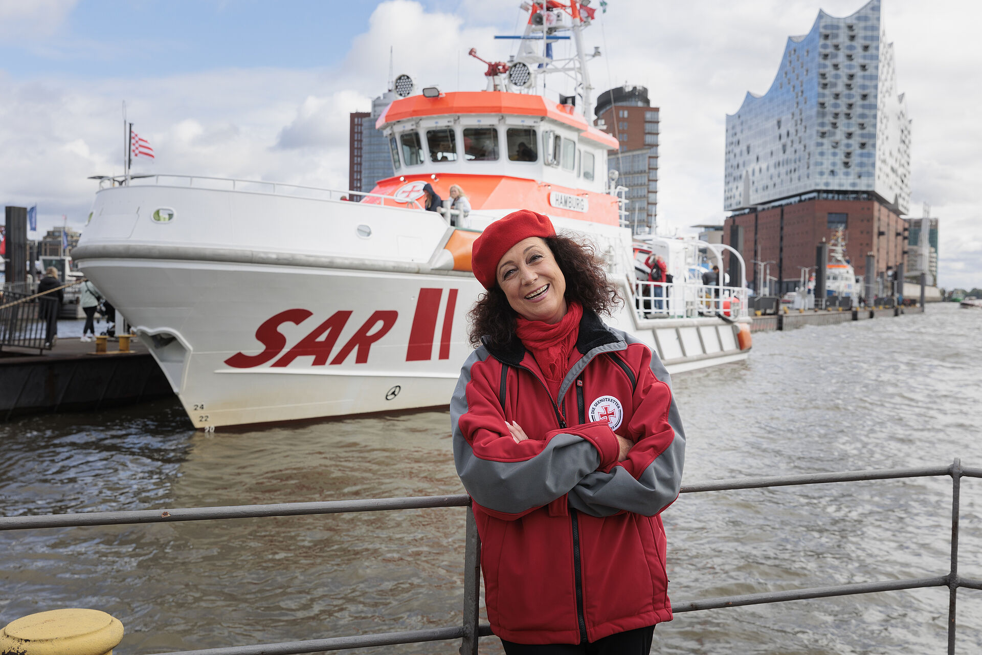 Die Schauspielerin Barbara Wussow steht vor einem rot-weißen Seenotrettungskreuzer, der am Hochhaus der Elbphilharmonie im Hamburger Hafen festgemacht hat.