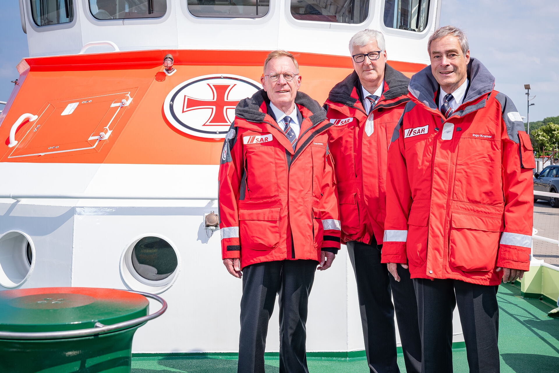 3 Herren in roter Wetterjacken stehen auf dem Vorschiff eines Seenotrettungskreuzer.