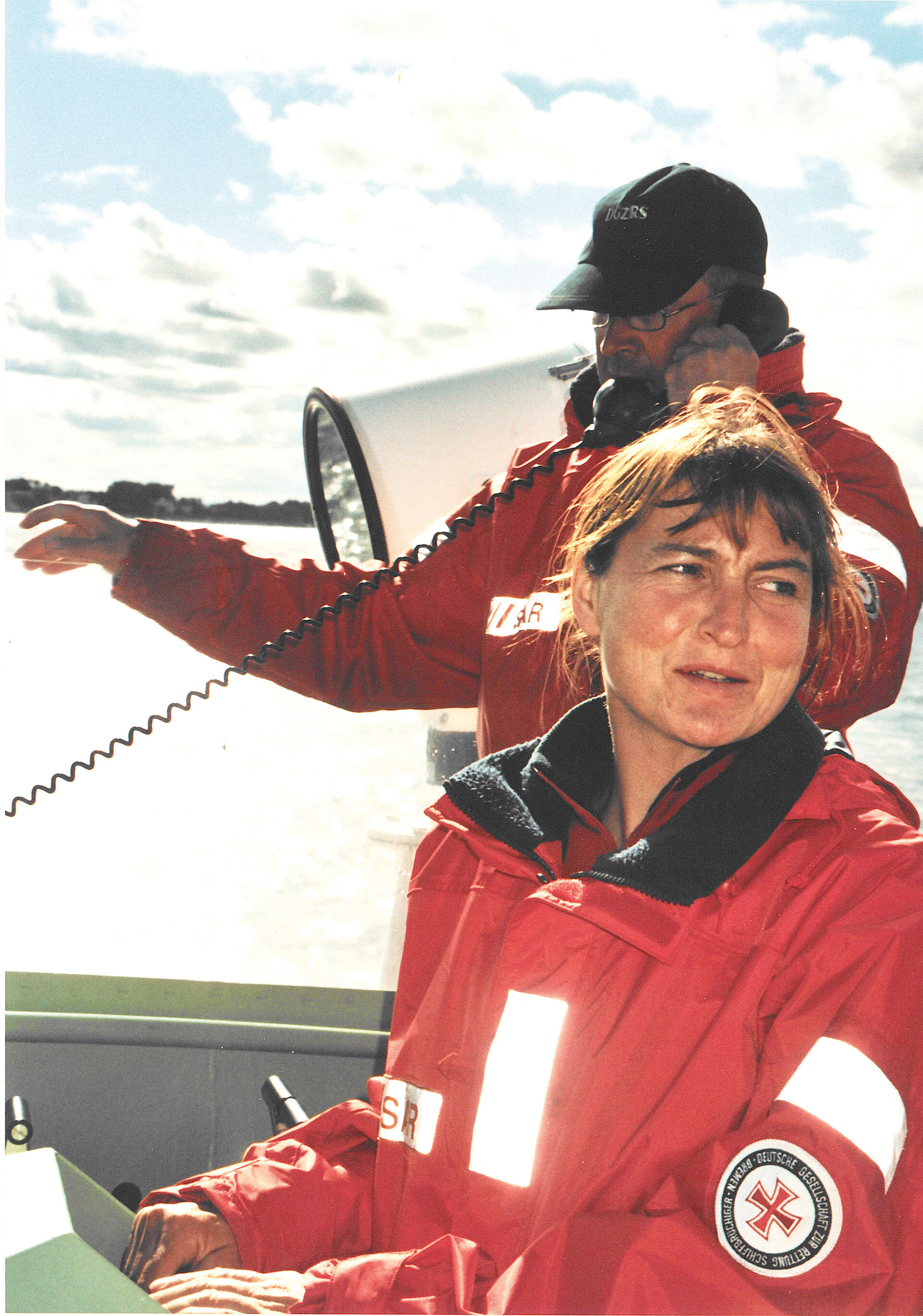 Eine Seenotretterin weist mit ihrem Finger an Deck in Richtung des Wassers, währen ein Seenotretter im Hintergrund einen Funkspruch abgibt