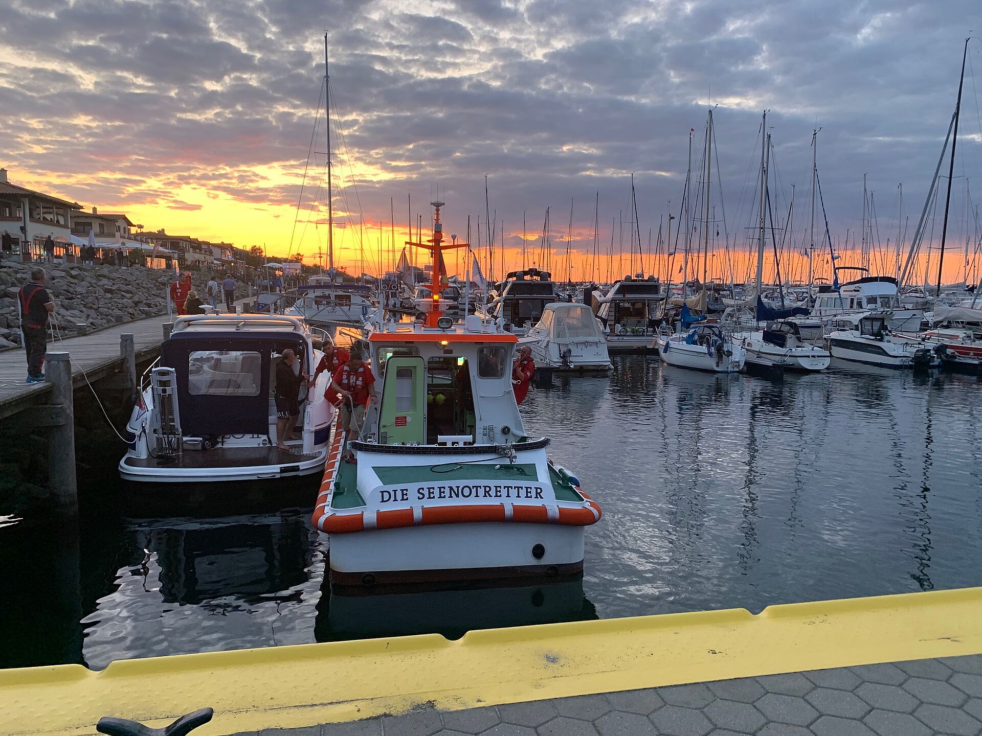 Im Sonnenuntergang liegt das Seenotrettungsboot neben einem kleinen Motorboot im Hafen von Kühlungsborn. Seenotretter helfen beim Festmachen und übernehmen Leinen.