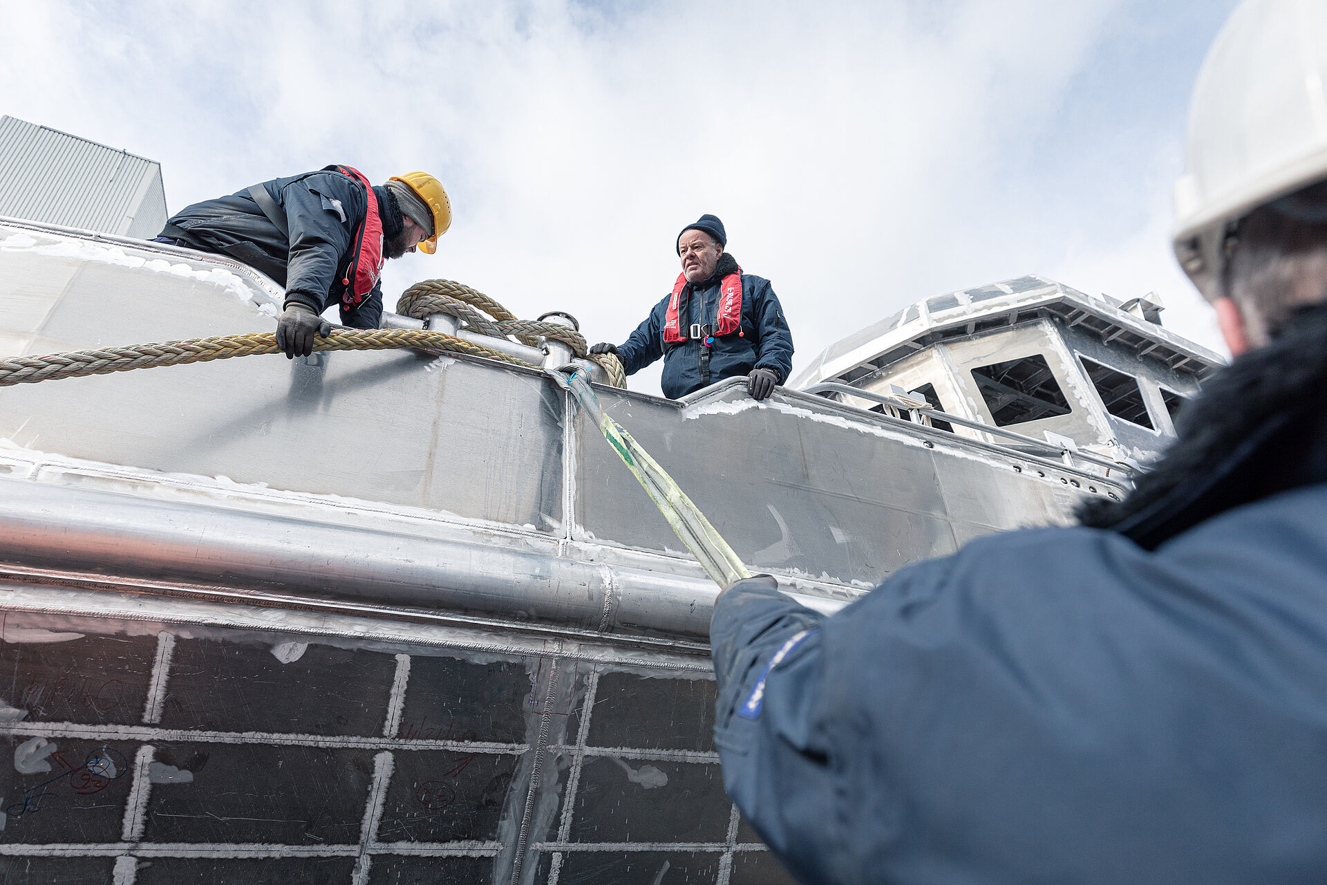 Blick über die Schulter eines Werftarbeiters auf den Aluminium-Rohbau eines Seenotrettungskreuzers. Auf dem Vorschiff des Rohbaus machen zwei Werftarbeiter dicke Festmacherleinen an einem Poller fest.