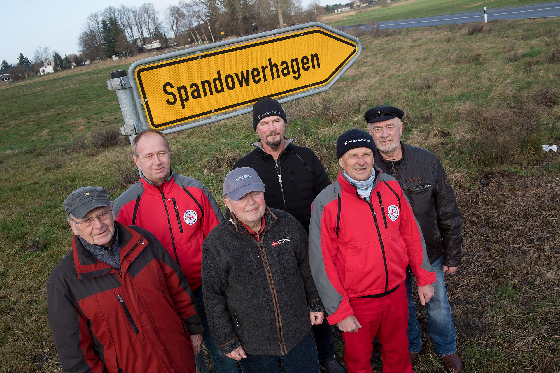 Eine Gruppe von 6 Männern steht vor dem gelben Verkehrschild mit der Aufschrift "Spandowerhagen". Zwei der Männer tragen rote Seenotretter-Kleidung.