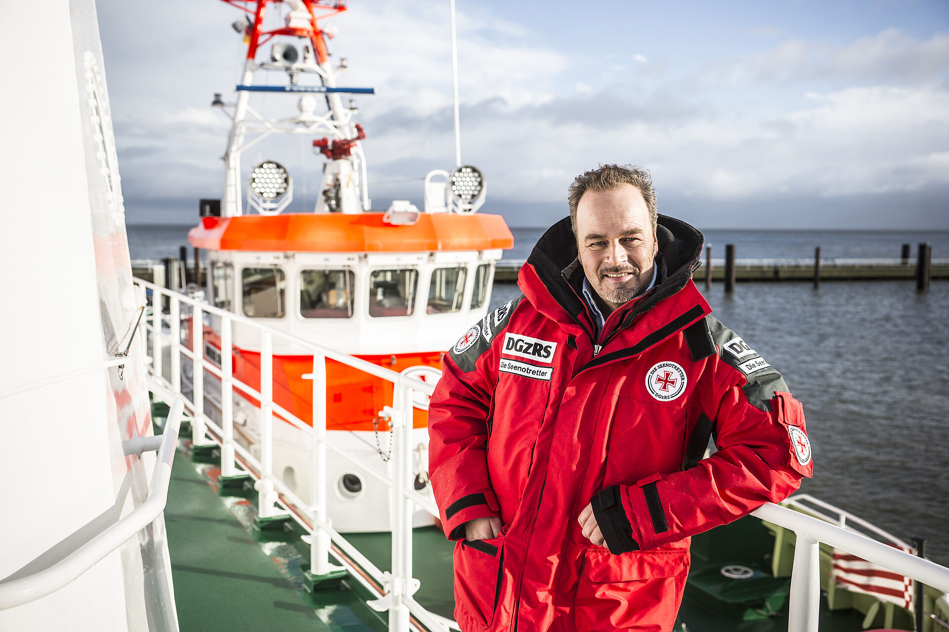 Der Schauspieler und Seenotretter-Botschafter Till Demtrøder lehnt in einer roten Wetterjacke der DGzRS auf einer Rehling. Im Hintergrund ist der Aufbau eines Seenotrettungskreuzers zu erkennen.
