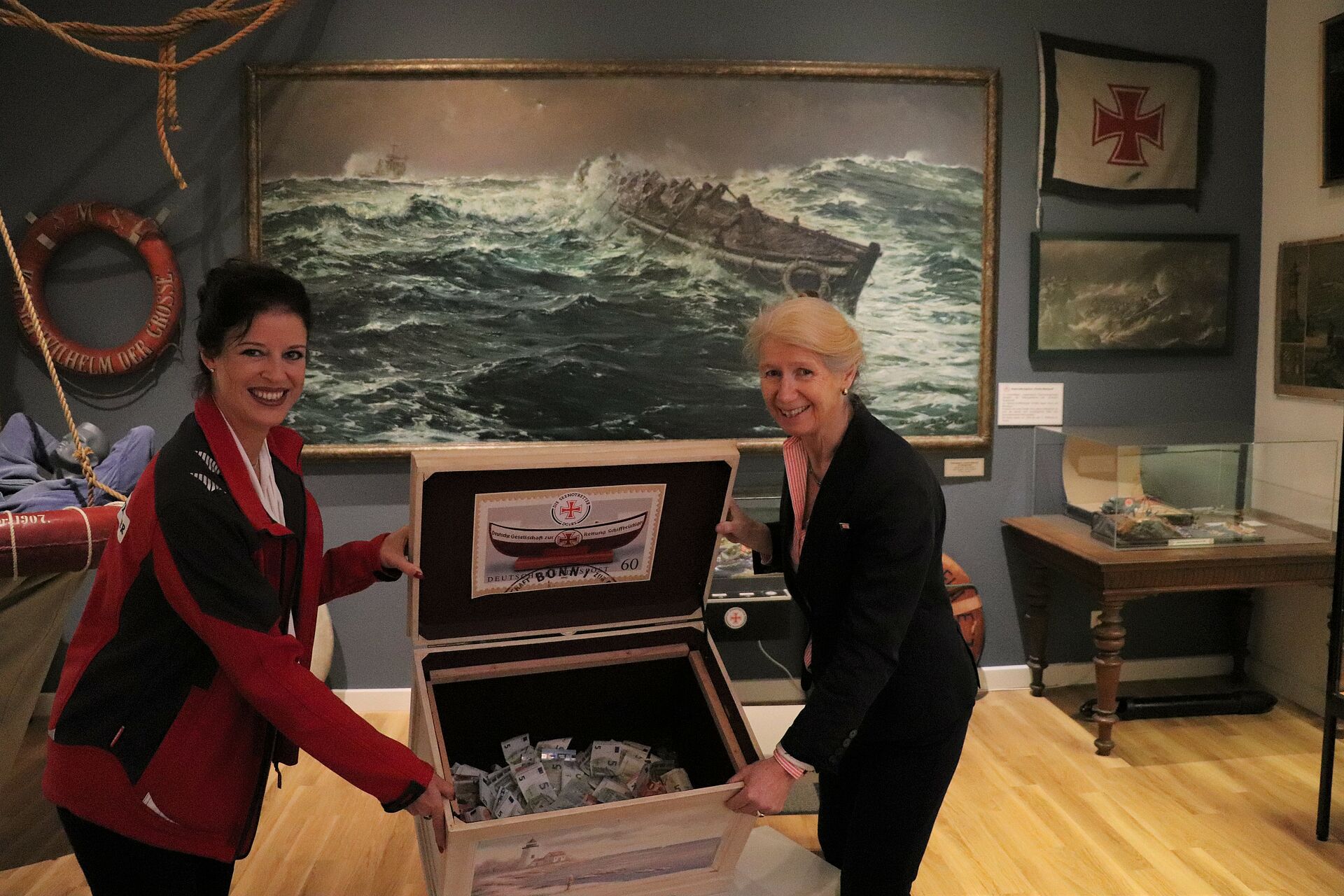 DGzRS-Mitarbeiterin Manuela Diestelhorst (l.) und Astrid Cohrs-Dreessen, Förderkreisvorsitzende des Schiffahrtsmuseums Nordfriesland, präsentieren in den Räumen der DGzRS-Ausstellung die zwischen ihnen stehende große Seemannskiste, die mit einer Spende an die Seenotretter gefüllt ist.