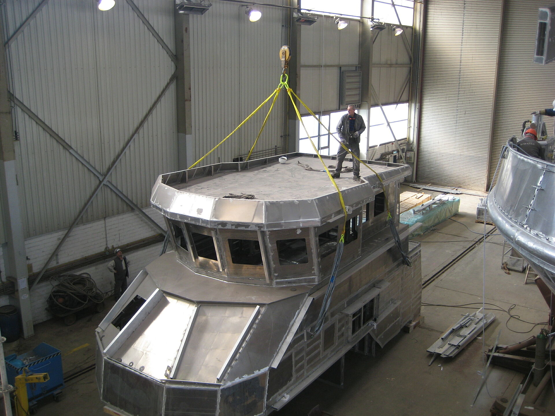 Ein Werftmitarbeiter steht auf dem Rohbau des Deckshauses aus Aluminium eines Seenotrettungskreuzers und befestigt gelbe Halteleinen des Hallenkranes.