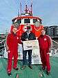 An Bord eines Seenotrettungskreuzers nehmen der Vormann (r.) und ein Seenotretter (l.) der DGzRS-Station Bremerhaven, beide in roter Dienstkleidung, einen symbolischen Spendenscheck vom Geschäftsführer der WindMW Service GmbH entgegen.  