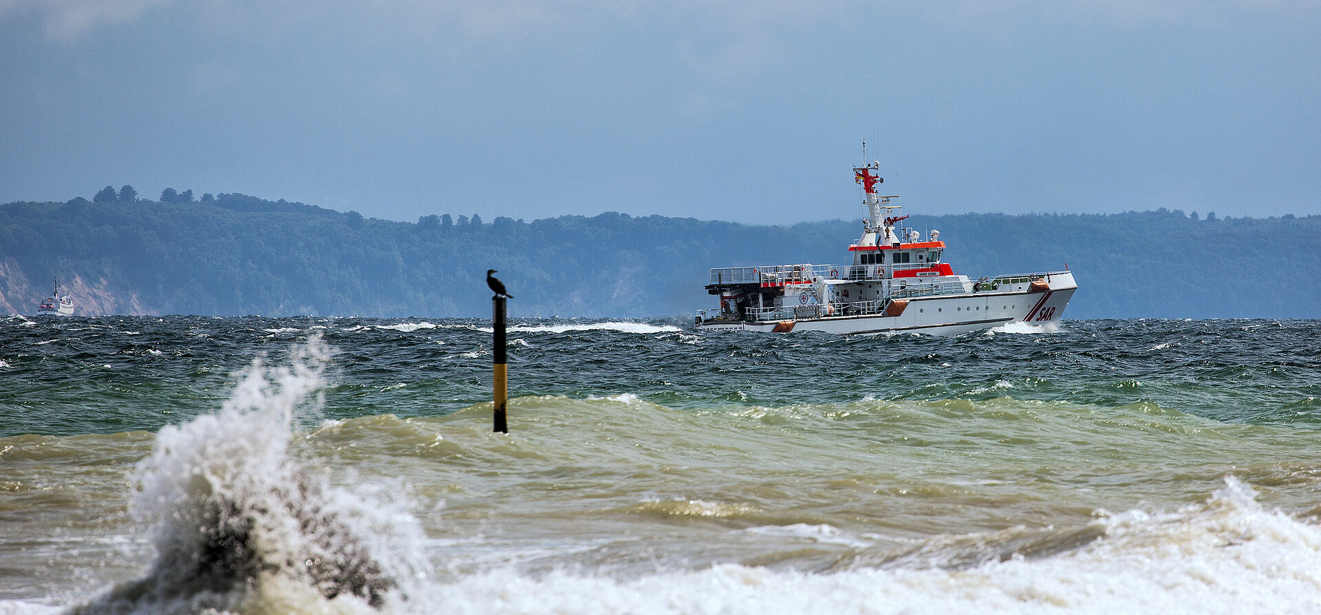 Der Seenotrettungskreuzer HARRO KOEBKE fährt auf der Ostsee vor der Silhouette der Insel Rügen. Im Vordergrund schlagen Wellen Gischt.