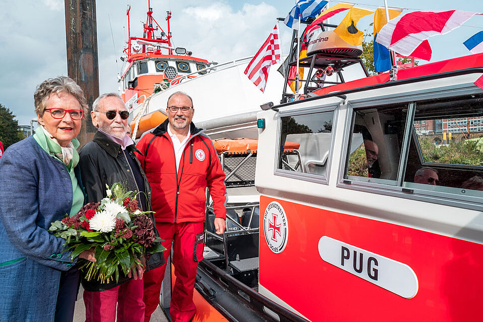 2 Männer und eine Frau stehen mit Blumenstrauß neben dem neu getauften Seenotrettungsboot PUG