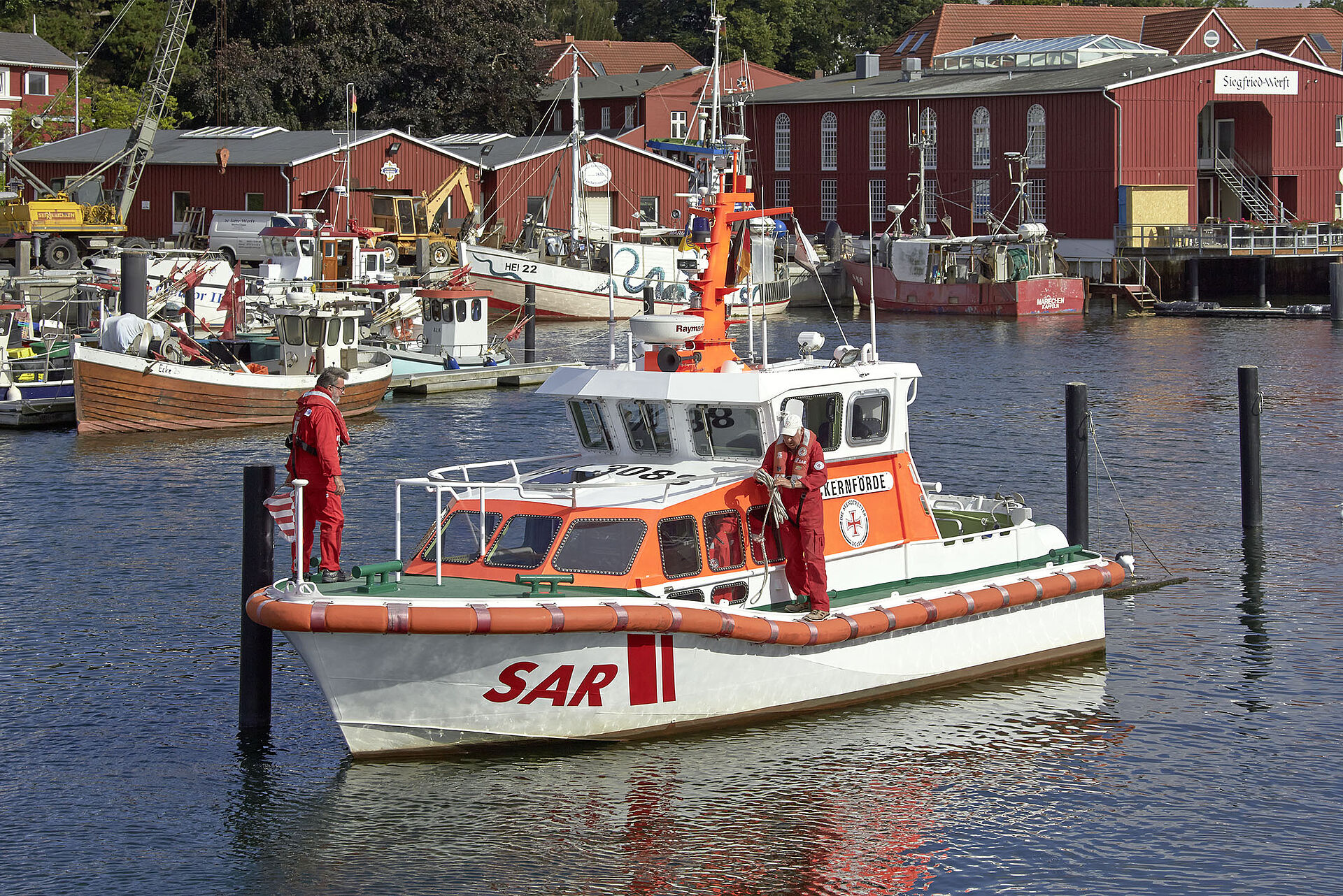 Im Hafenbecken von Eckernfoerde rangiert bei gutem Wetter das Seenotrettungsboot. Zwei Seenotretter machen Leinen klar.