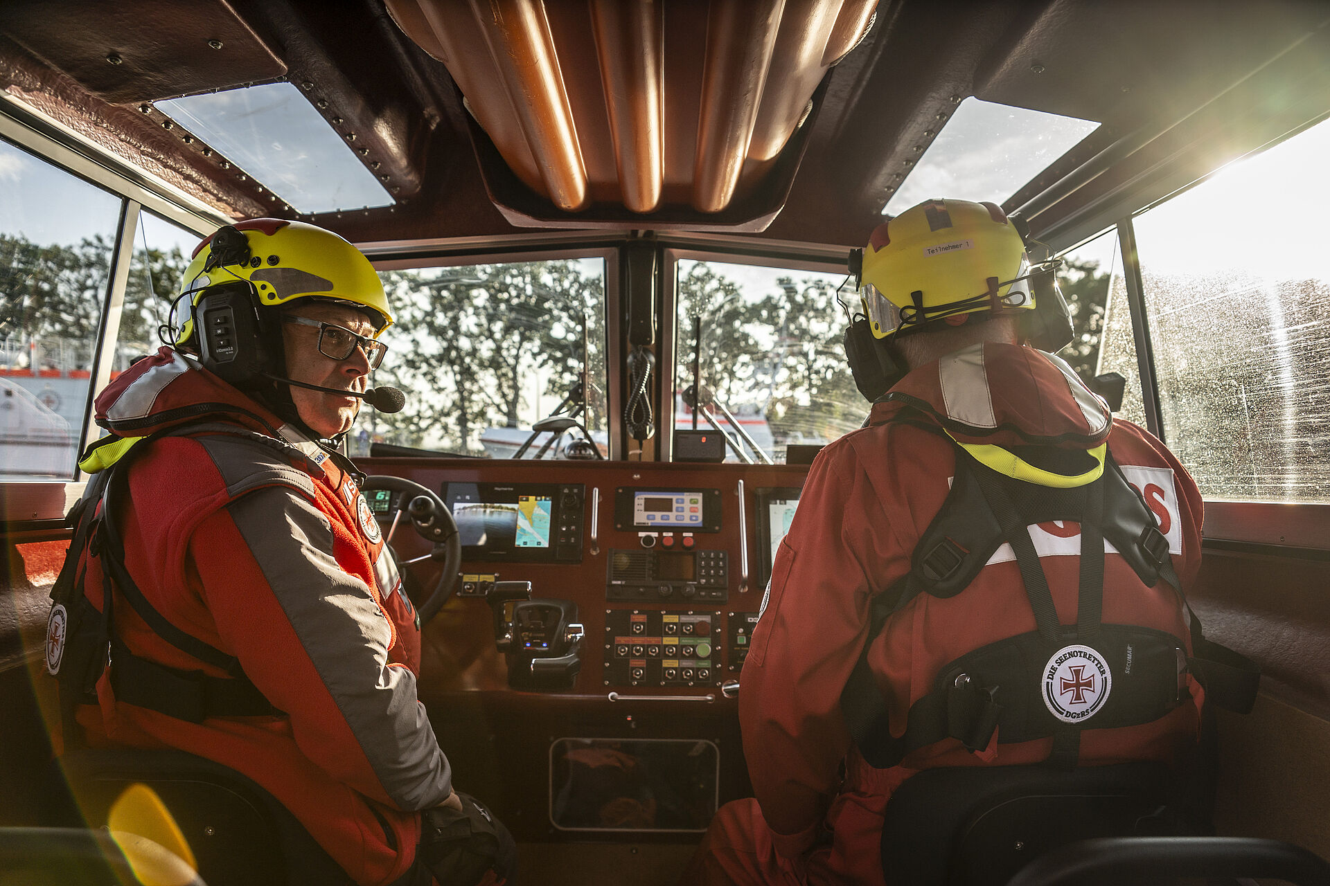 Zwei Seenotretter sitzen am Steuer eines Seenotrettungsbootes. Sie tragen eine Rettungsweste und einen Helm mit Headset.