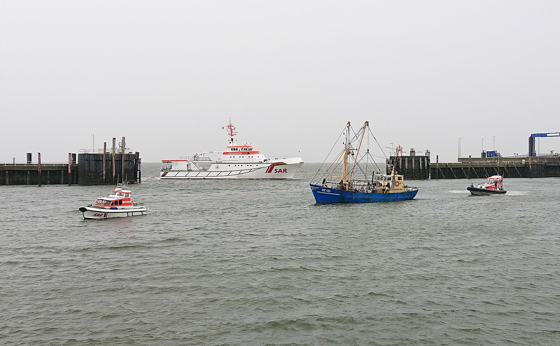 Zwei Tochterboote eines Seenotrettungskreuzer haben in einer Hafeneinfahrt einen Fischkutter in Schlepp genommen, der große Kreuzer liegt im Hintergrund