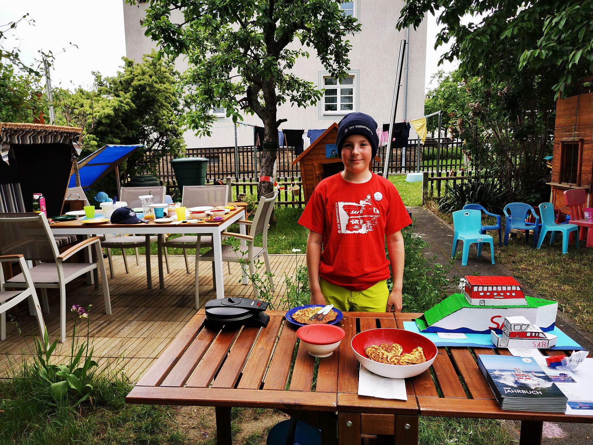 Ein Junge im roten Seenotretter-T-Shirt und blauer Seenotretter-Strickmütze steht in einem Hinterhofgarten. Vor ihm, auf einem Holztisch aufgebaut, selbstgebackene Waffeln sowie zwei selbstgebastelte Papiermodelle von Seenotrettungskreuzern und ein Stapel DGzRS-Jahrbücher. 