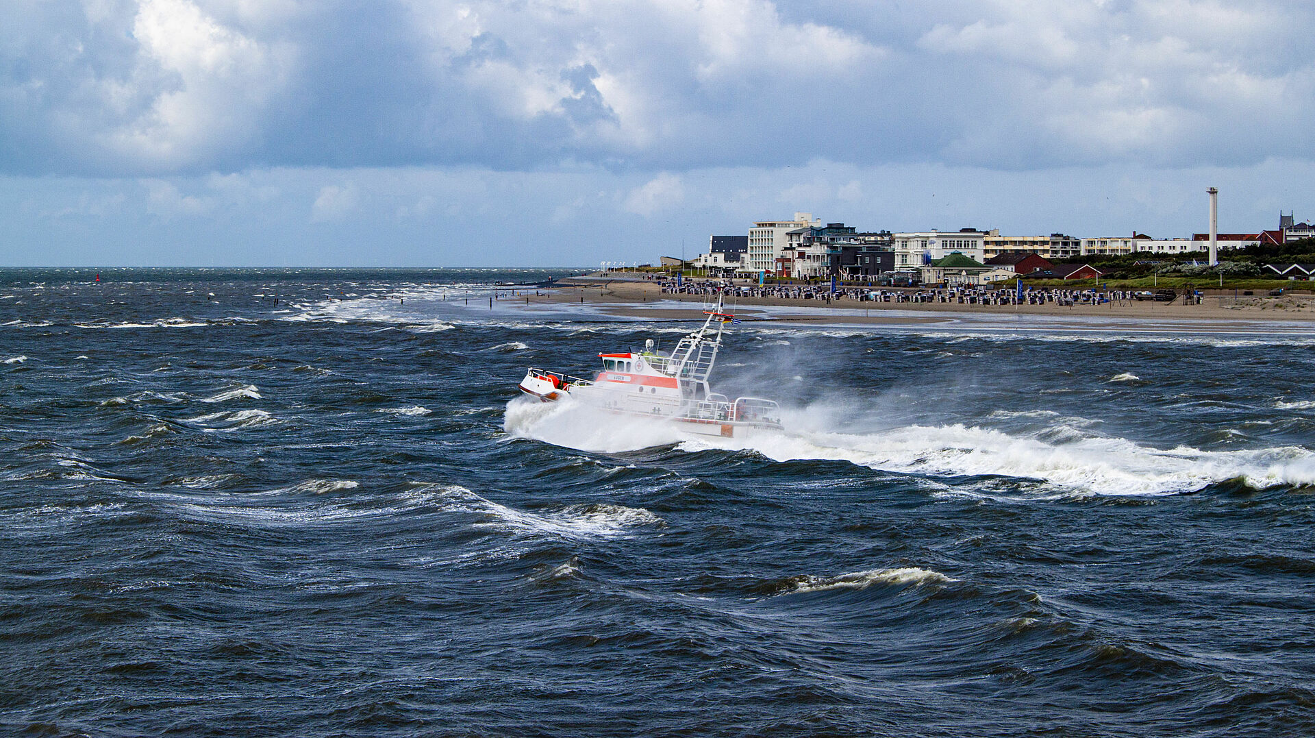 Vor der Kulisse des Norderneyer Strandes verlässt der Seenotrettungskreuzer EUGEN den Hafen Richtung offene Nordsee. Die Wellen schlagen Gischt.
