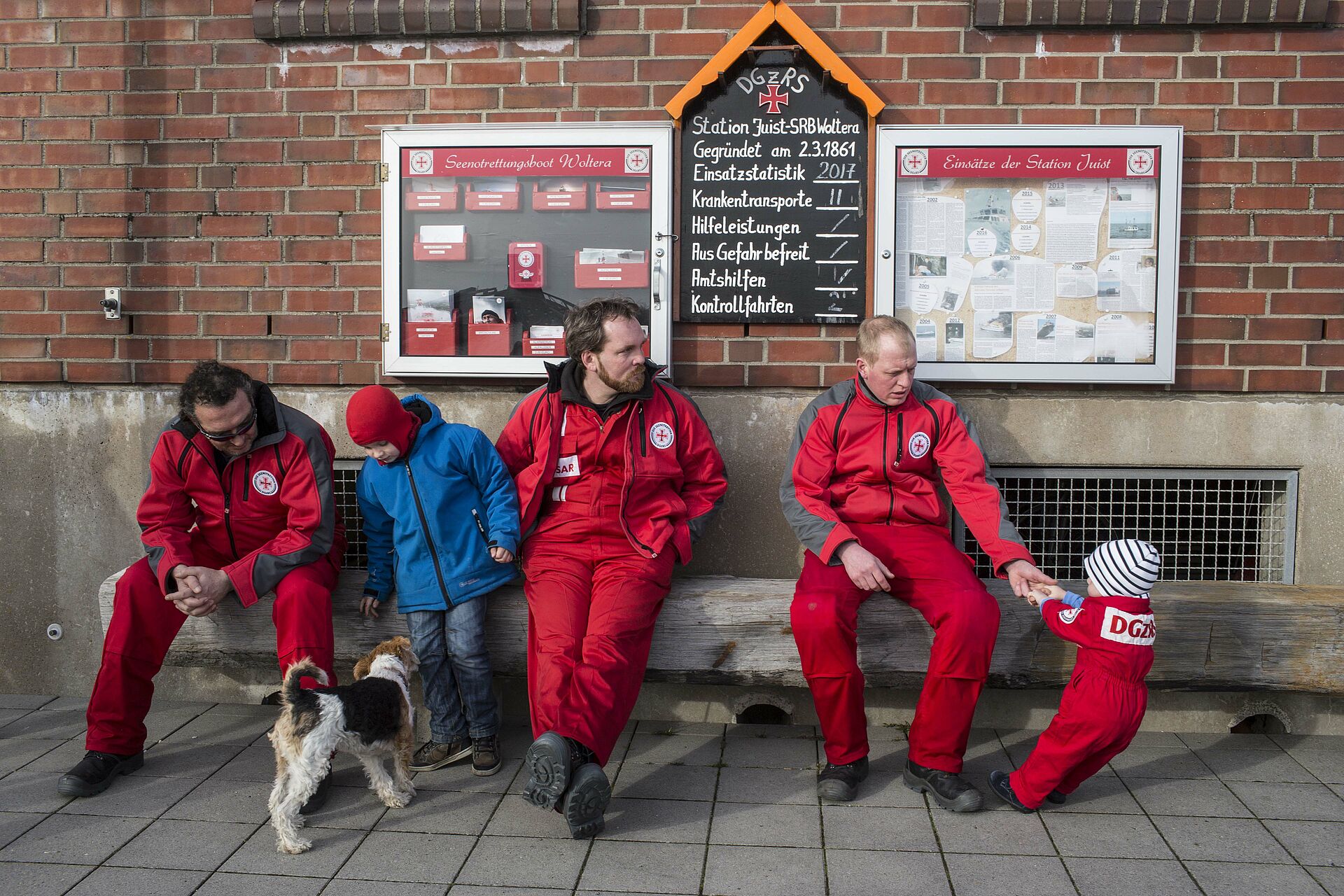 Drei Seenotretter, mit einem Hund und zwei Kindern, sitzen auf der Bank vor einem Stationsgebäude. 