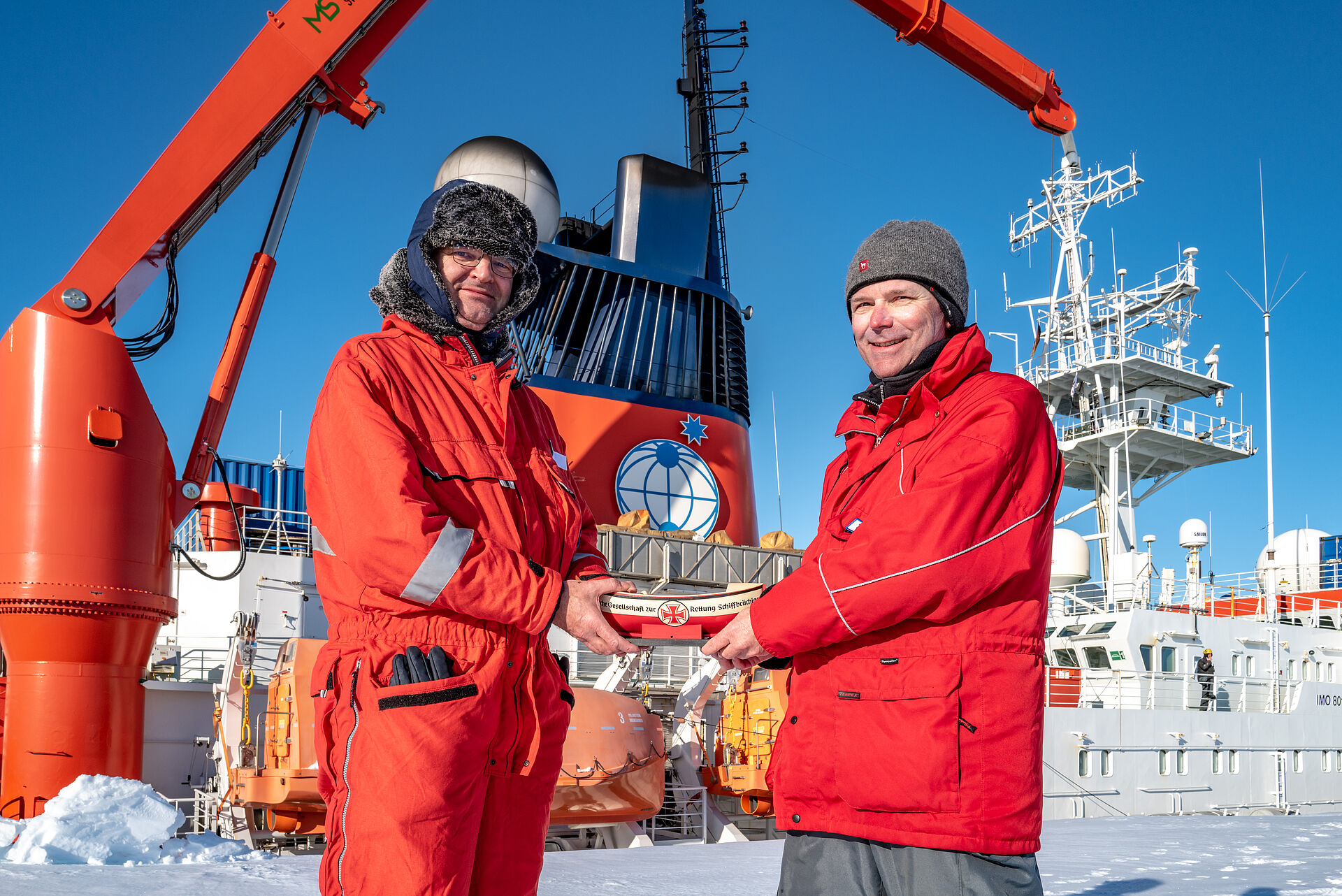 In der Antarktis überreicht vor dem Forschungsschiff „Polarstern“ ein Mann einem anderen Mann ein Sammelschiffchen der DGzRS.