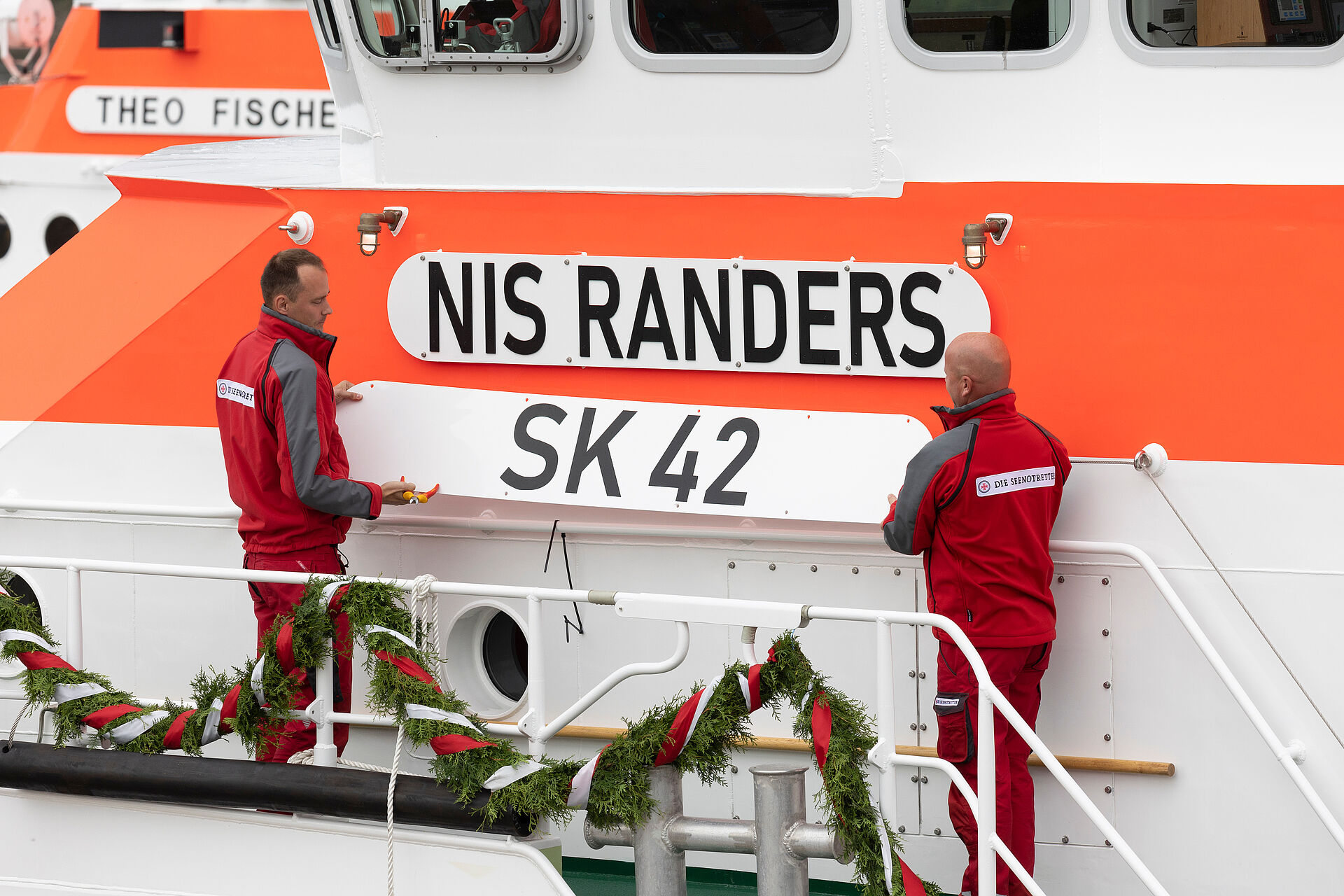 Zwei Seenotretter stehen am Seitengang des Seenotrettungskreuzers und heben das Schild mit der Beschriftung SK 42 runter. Dahinter hängt das Schild mit der Beschriftung NIS RANDERS.