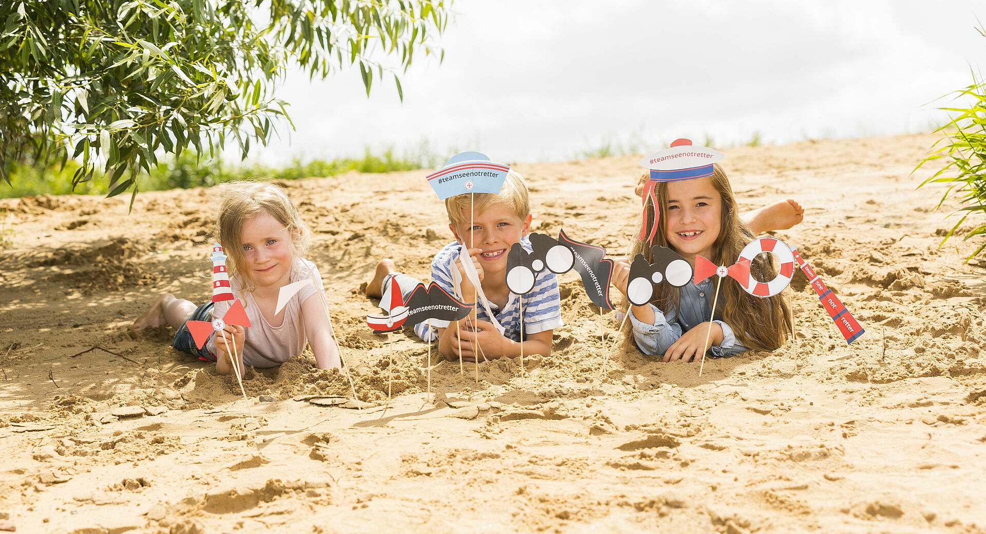 Drei Kinder liegen mit selbstgebastelten DGzRS Material im Sand
