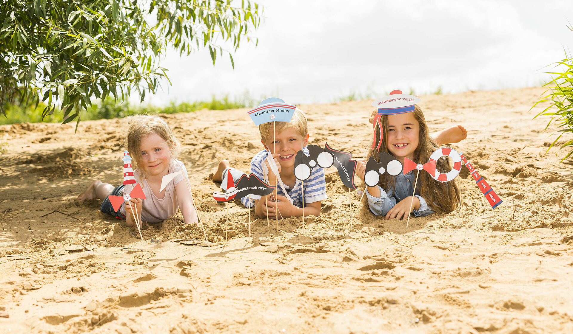 Drei Kinder liegen mit selbstgebastelten DGzRS Material im Sand