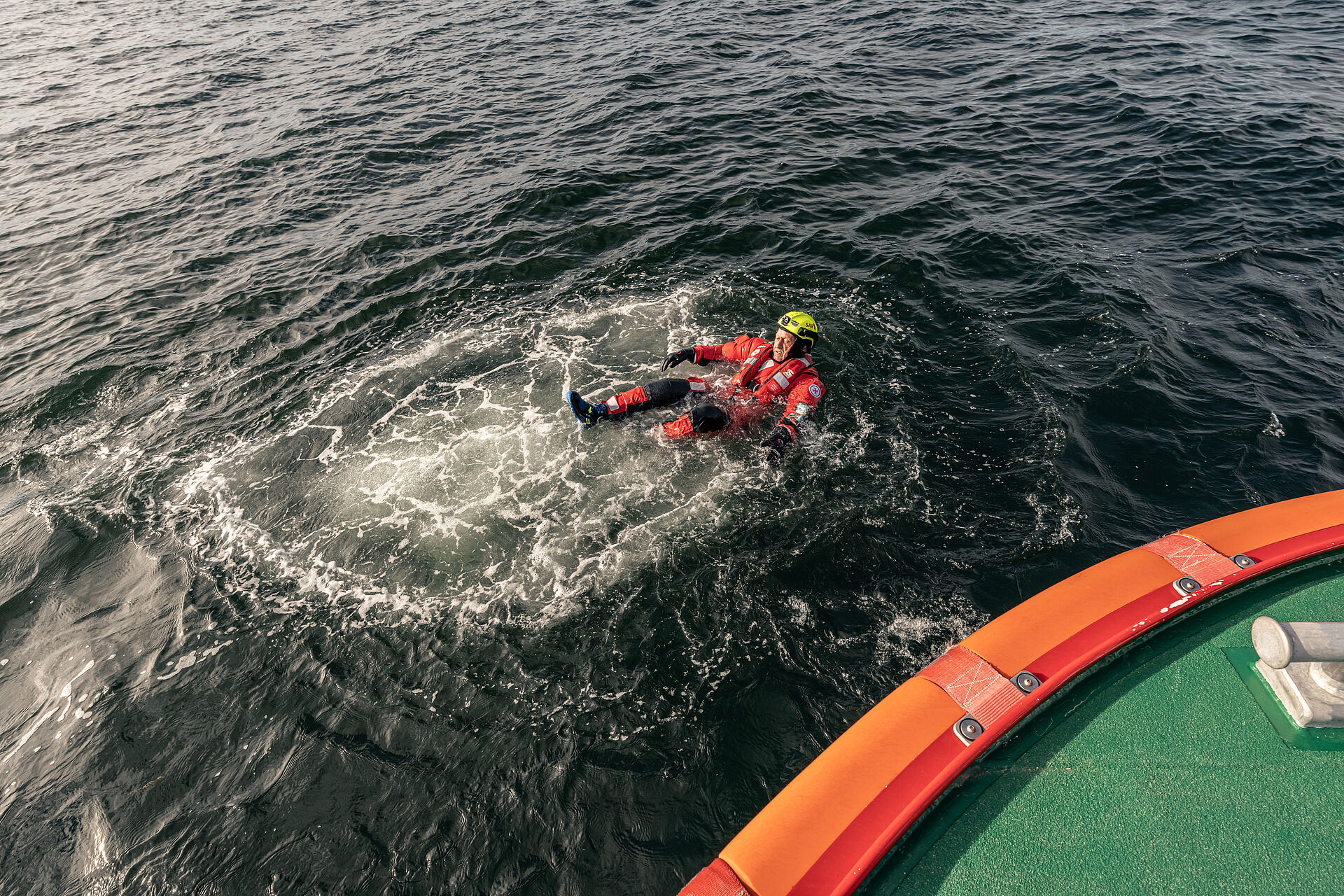 Ein Seenotretter ist in Überlebensanzug ins Wasser gesprungen und treibt auf dem Rücken liegend neben dem Seenotrettungsboot.