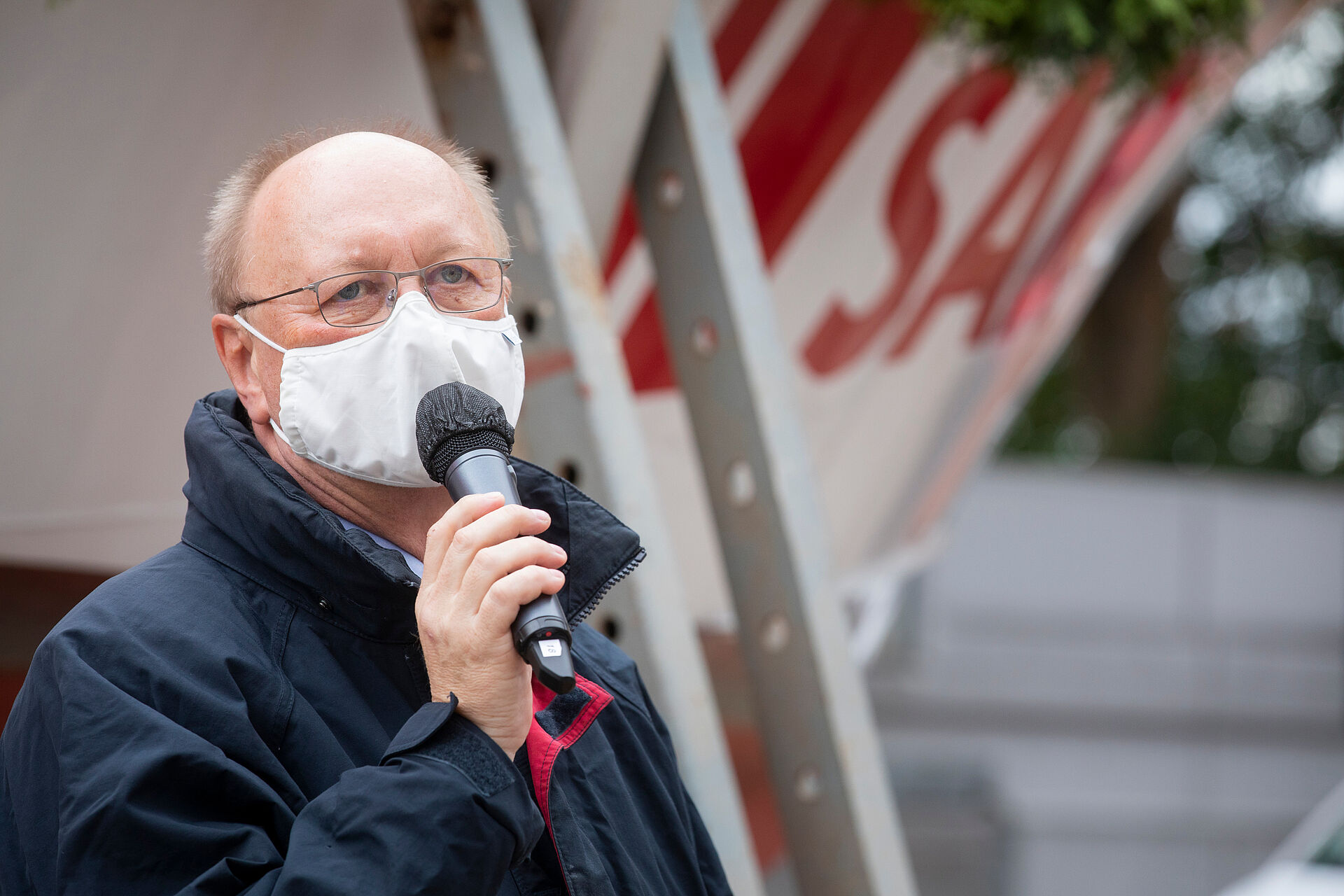 DGzRS-Geschäftsführer Michael Ippich spricht in ein Mikrofon, das er in der Hand hält. Er trägt einen Mund-Nasen-Schutz und steht vor dem Rumpf des neuen Seenotrettungsbootes.