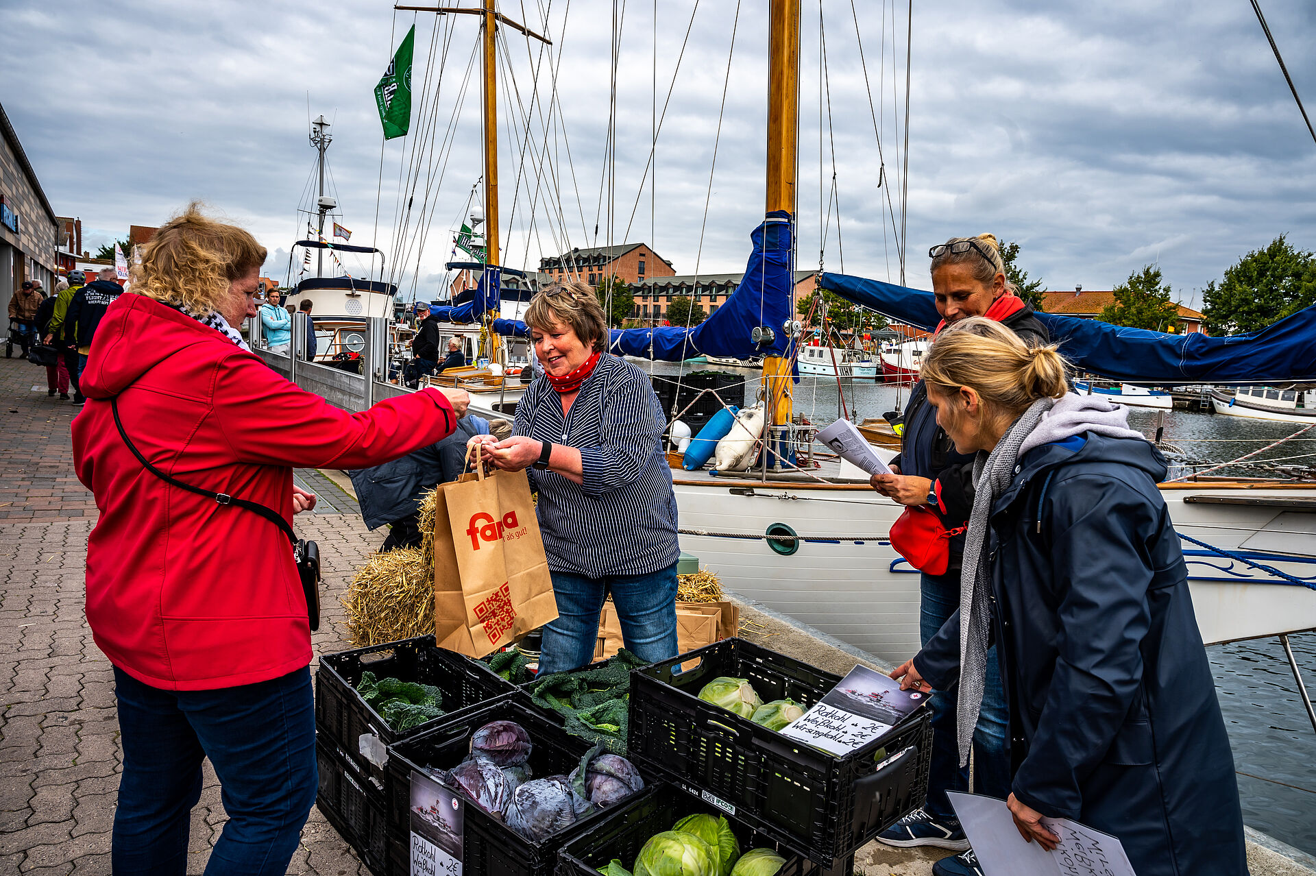 Verkauf von Kohlköpfen im Hafen von Heiligenhafen. Eine Verkäuferin übergibt eine Tüte mit dem Gemüse an eine Kundin, zwei weitere Damen stehen an und schauen das Angebot an. 