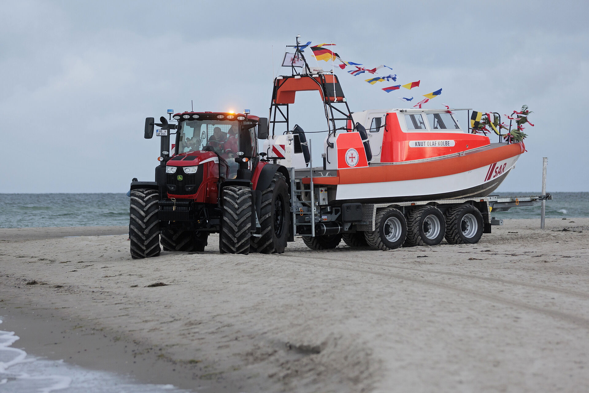 Ein Traktor zieht einen Anhänger mit einem Seenotrettungsboot auf dem Strand zur Wasserkante.